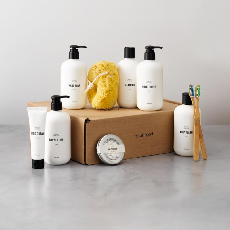 A selection of Public Goods products such as body wash, hand soap, shampoo, and conditioner are displayed on and around a brown cardboard box with the words, "it's all good" printed on the side. The products all have white packaging with black text. Among the packaged products is a shower sponge and two wooden toothbrushes.